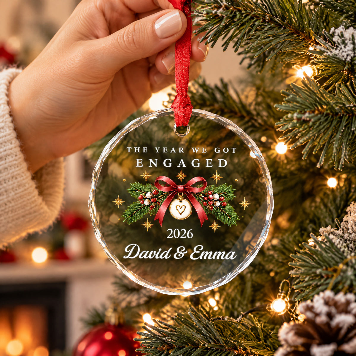 Personalized Glass Ornament Engaged Couple held by a hand in a knit sleeve beside a tree with warm lights