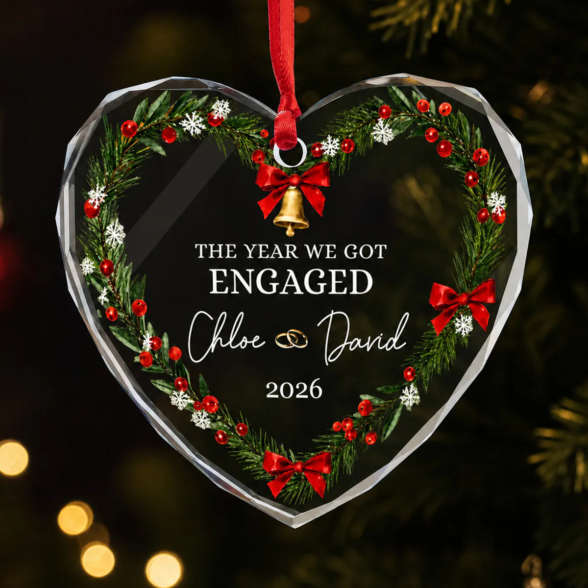 Personalized glass ornament heart with wreath, red bows and bell, engaged text, hanging on a dark tree
