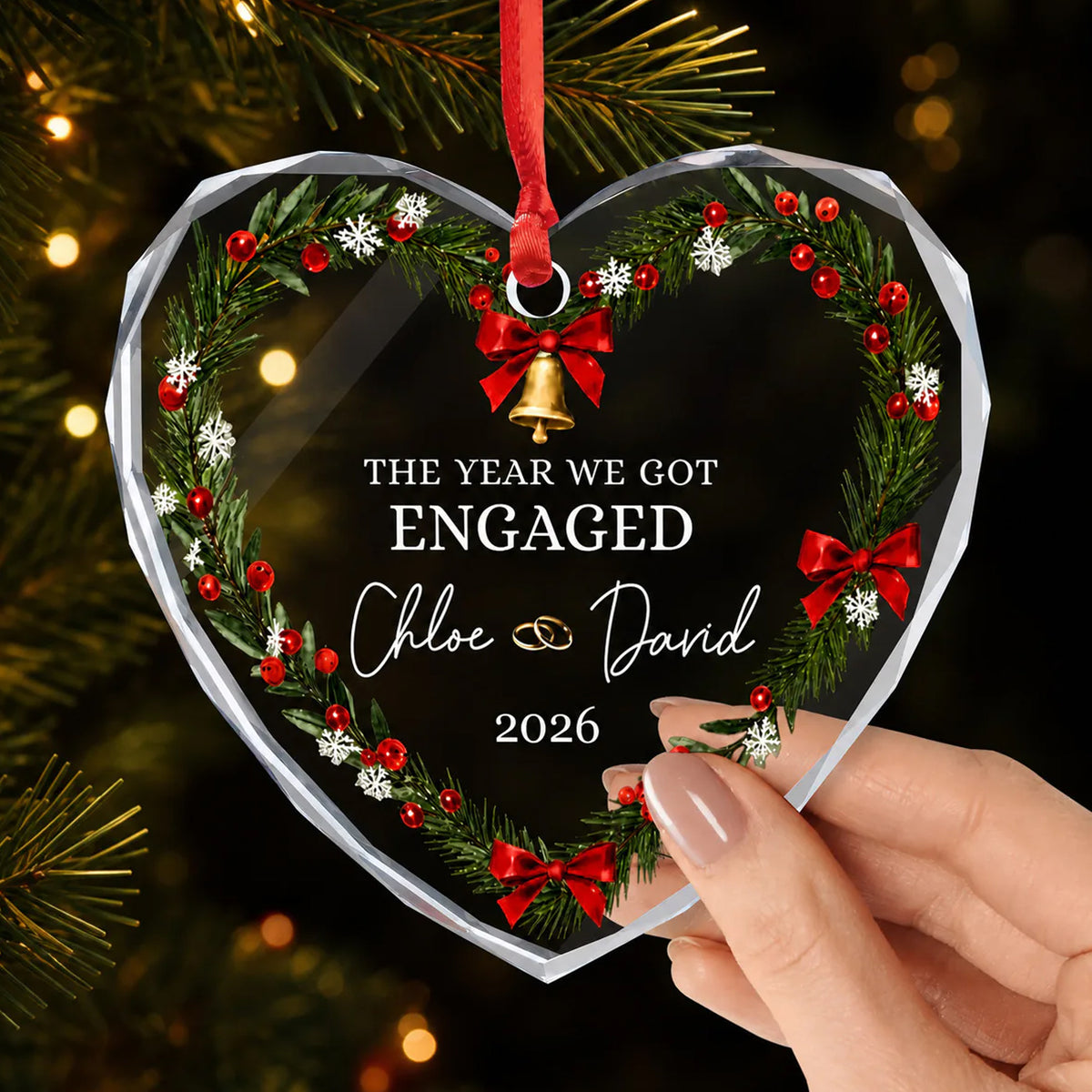 Personalized glass ornament held in a hand near pine branches and warm lights, showing names and 2026