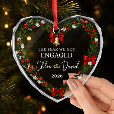 Personalized glass ornament heart with wreath, red bows and bell, engaged text, hanging on a dark tree