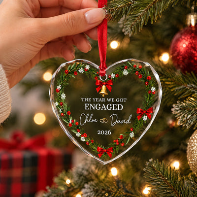 Personalized glass ornament held by a hand on a Christmas tree beside red baubles and a plaid gift