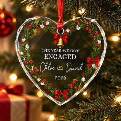 Personalized glass ornament hanging from red ribbon on a lit tree with a wrapped gift in the background