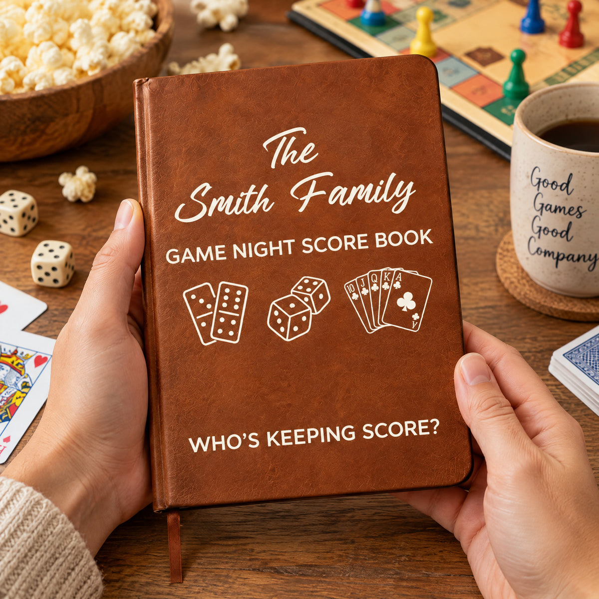 Personalized leather journal game night score held open-handed beside popcorn, a board game, dice and a mug on a coaster