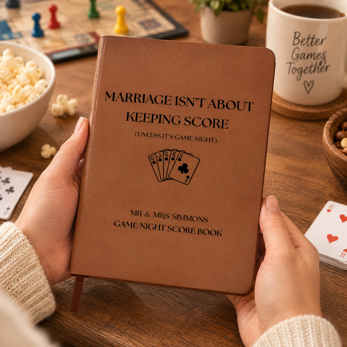 Personalized leather journal score book held over a table with popcorn, board game pieces, a mug and playing cards