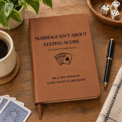 Personalized leather journal score book on a wooden table with coffee cup, plant, dice, gold-trimmed pen and playing cards