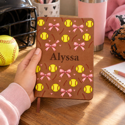 Personalized softball pattern leather journal held in one hand near a softball, helmet and glove on a desk