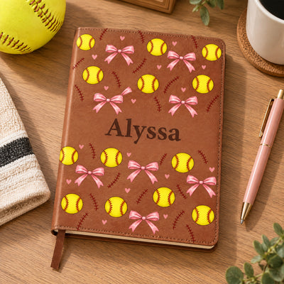 Personalized softball pattern leather journal on a wood table with a pink pen, softball and towel nearby