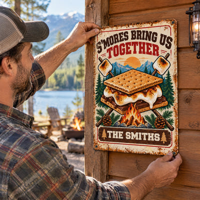 Personalized metal sign S'mores Bring Us Together held against a wood cabin wall by a man in a plaid shirt