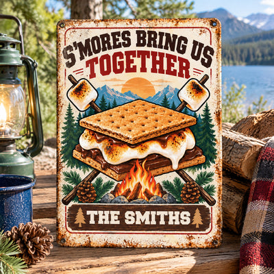 Personalized metal sign S'mores Bring Us Together on a wooden table beside a lantern, firewood and plaid blanket