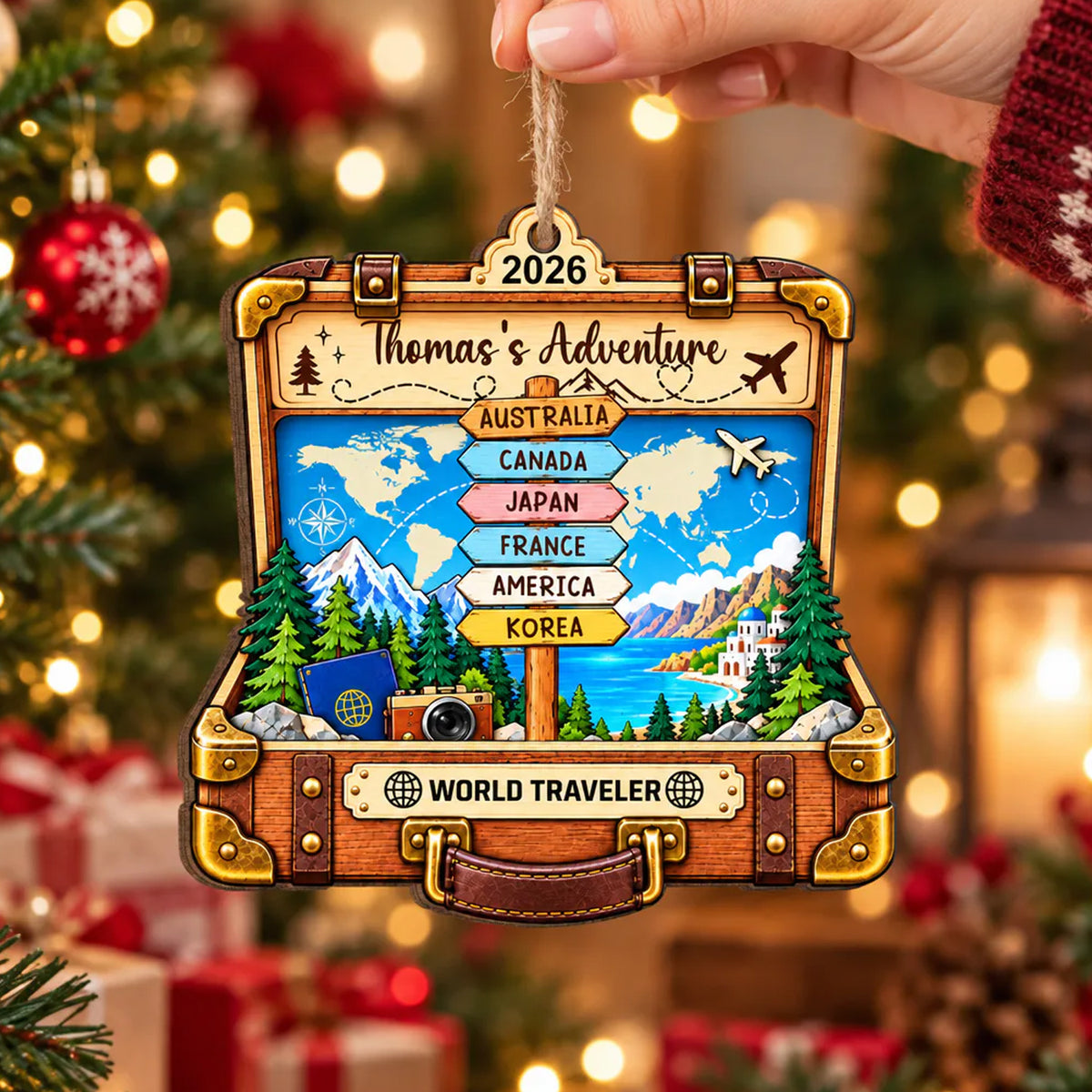 Personalized suitcase wooden ornament held by a hand in front of a lit Christmas tree with red baubles