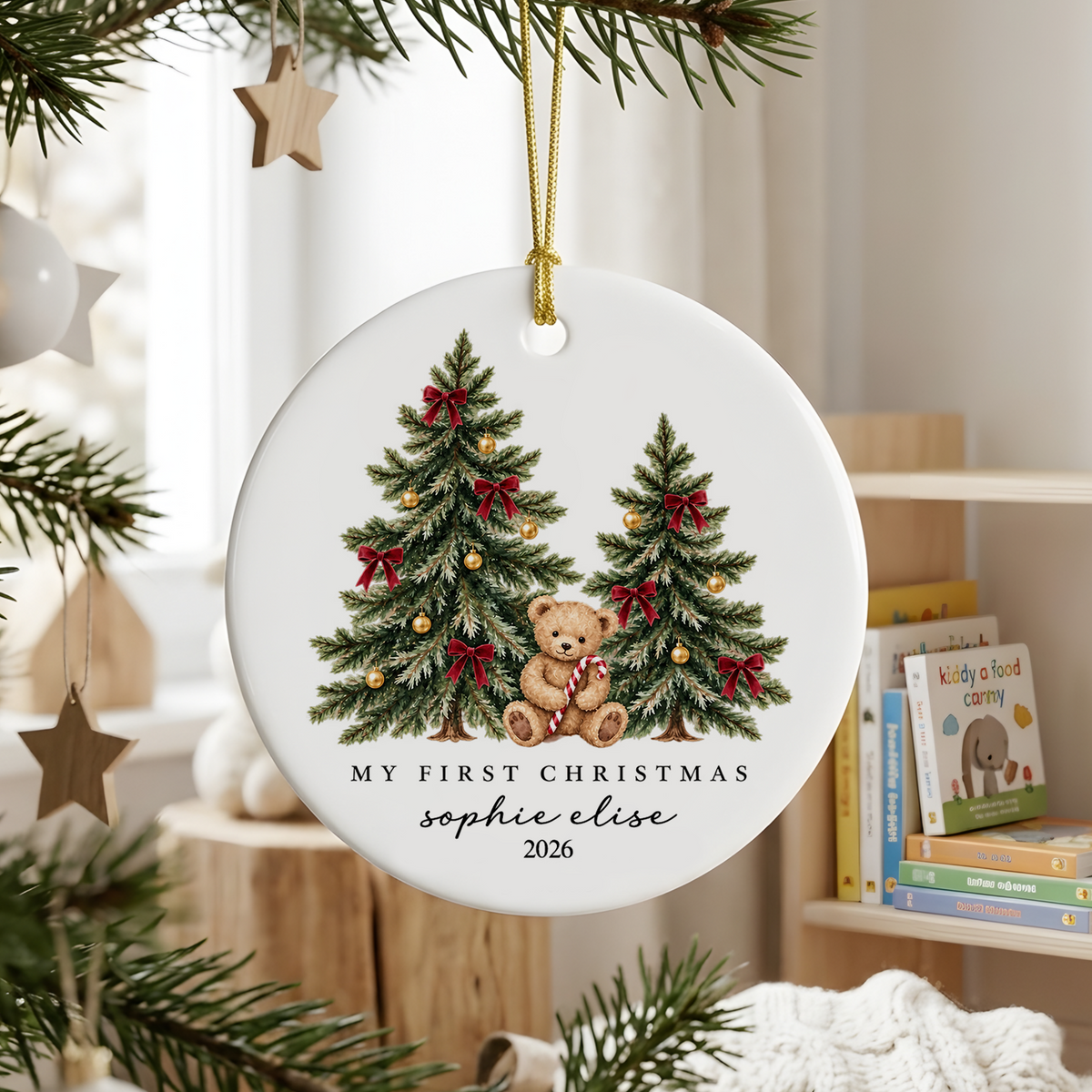Personalized ceramic Christmas ornament teddy bear hanging on a pine branch near a window and children's books