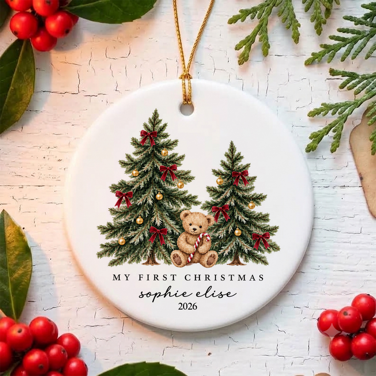 Personalized ceramic Christmas ornament teddy bear flat on a white wood surface with red berries and greenery