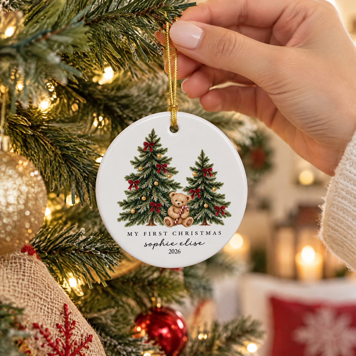 Personalized ceramic Christmas ornament teddy bear held by hand in front of a lit tree with warm glowing lights