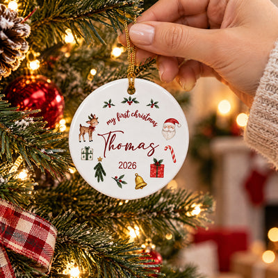 Personalized First Christmas Ceramic Ornament round, held by a hand near tree branches and a plaid ribbon