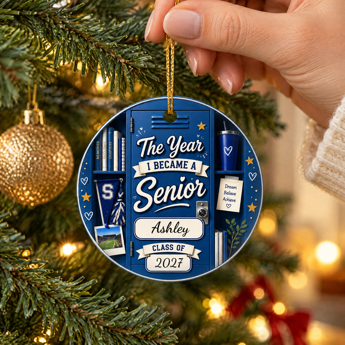 Personalized ceramic senior class ornament in blue held by hand on a Christmas tree beside a gold bauble