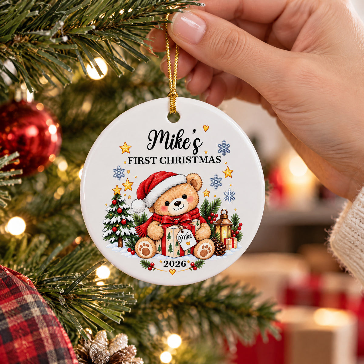 Personalized Ceramic Ornament First Christmas held by a hand in front of a tree with red baubles and a plaid fabric