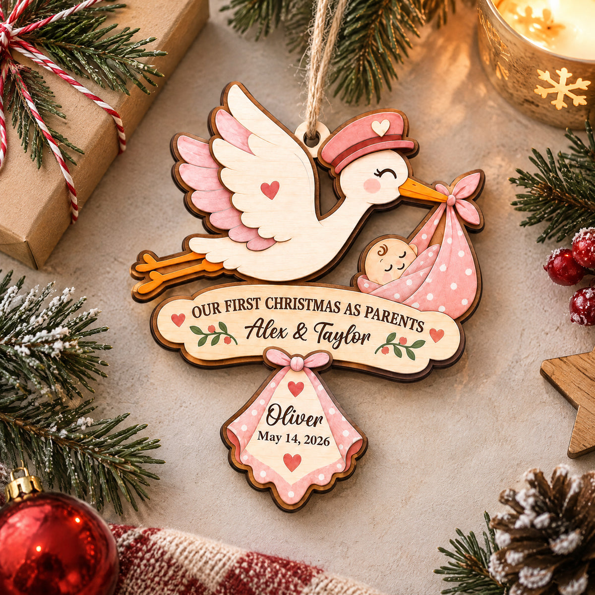 Our First Christmas As Parents Personalized Wooden Ornament, pink stork lying flat near gift, candle and pine sprigs