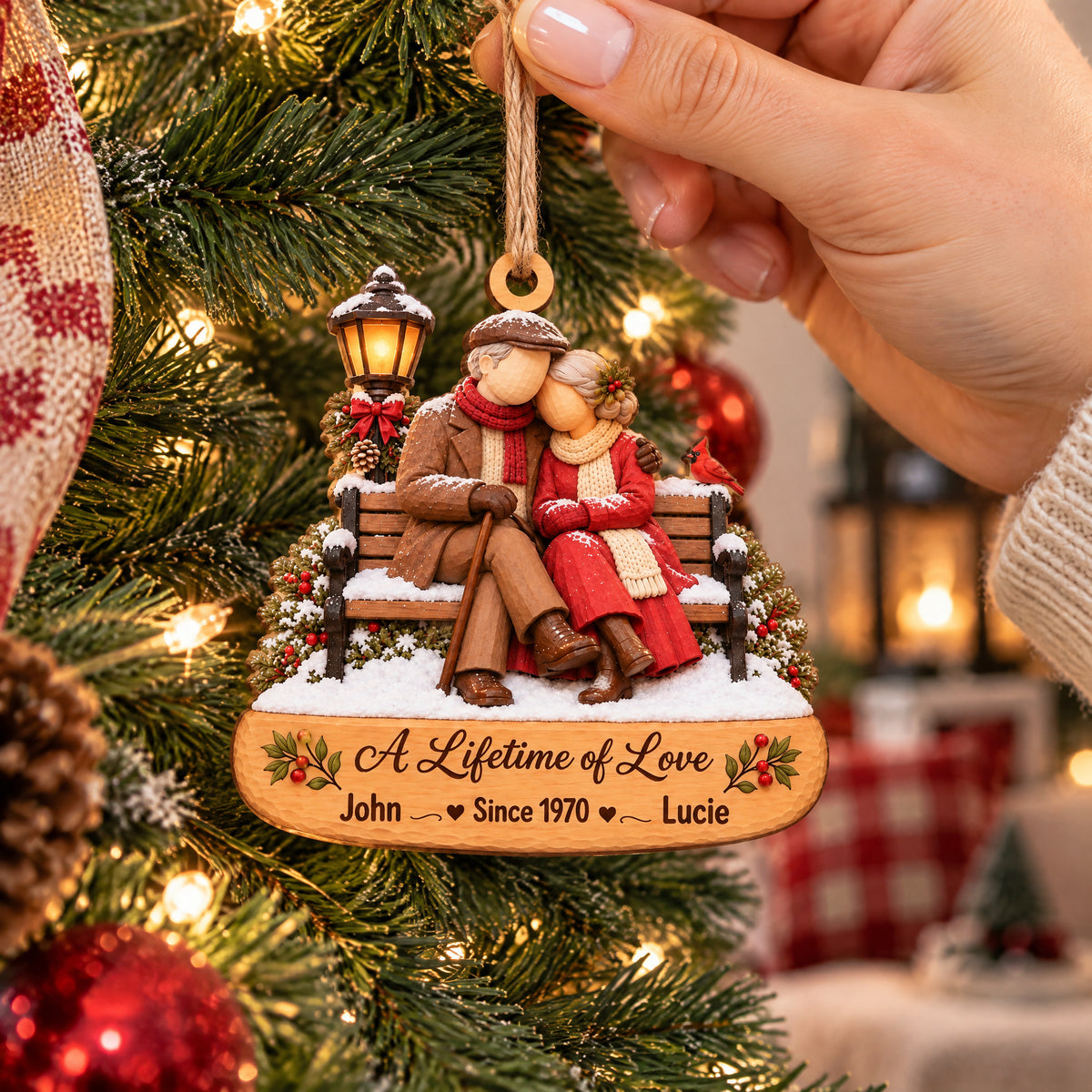 Personalized Wooden Couple Christmas Ornament held by a hand beside a lit tree, with red plaid and a pinecone nearby