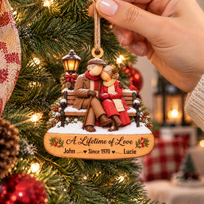 Personalized Wooden Couple Christmas Ornament held by a hand beside a lit tree, with red plaid and a pinecone nearby