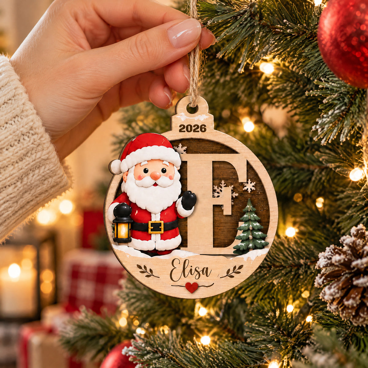 Personalized Santa Initial Wooden Ornament held by a hand in a knit sleeve beside tree lights and a pinecone