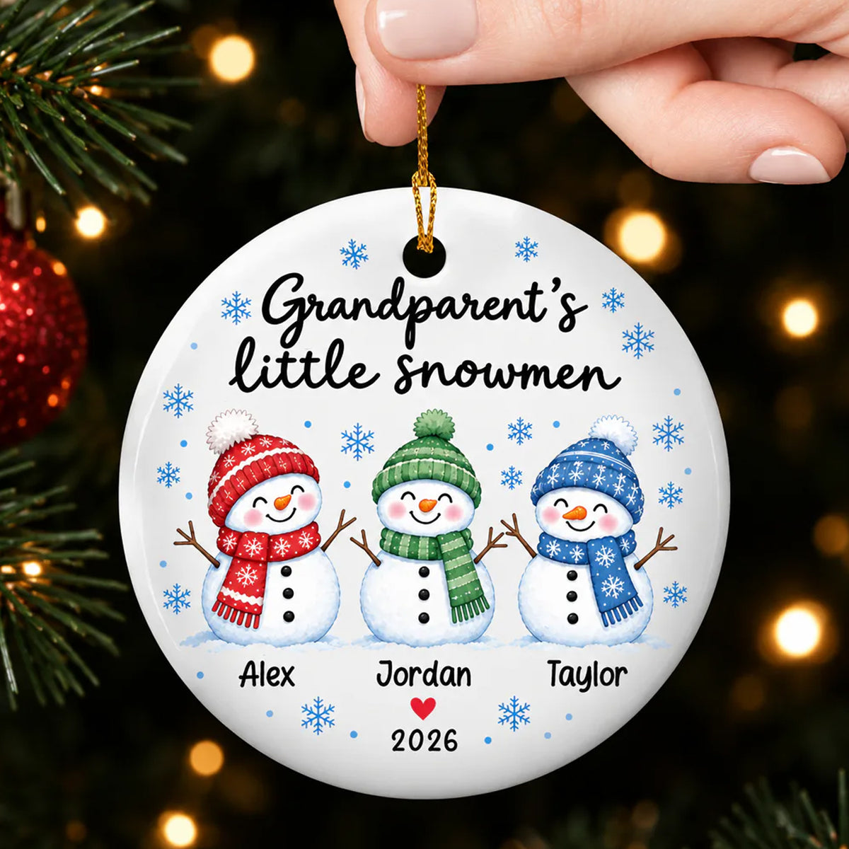 Personalized Grandparent's Little Snowmen Ceramic ornament held by a hand by its gold cord near tree lights