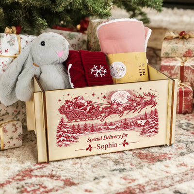 Personalized Christmas Eve Box filled with a gray bunny, red knit socks and pink blanket under a tree beside wrapped gifts