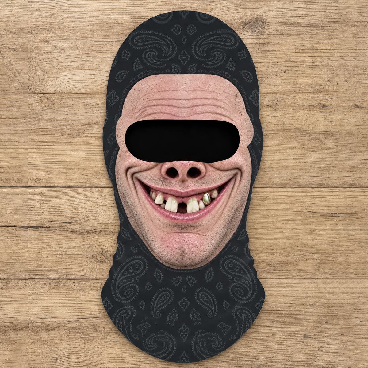 Funny Personalized Ski Mask laid flat on a wood surface, black paisley fabric with a printed grinning face