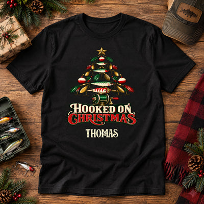 Hooked On Christmas Personalized Shirt in black laid flat on wood with lures, pinecones, a plaid scarf and a cap