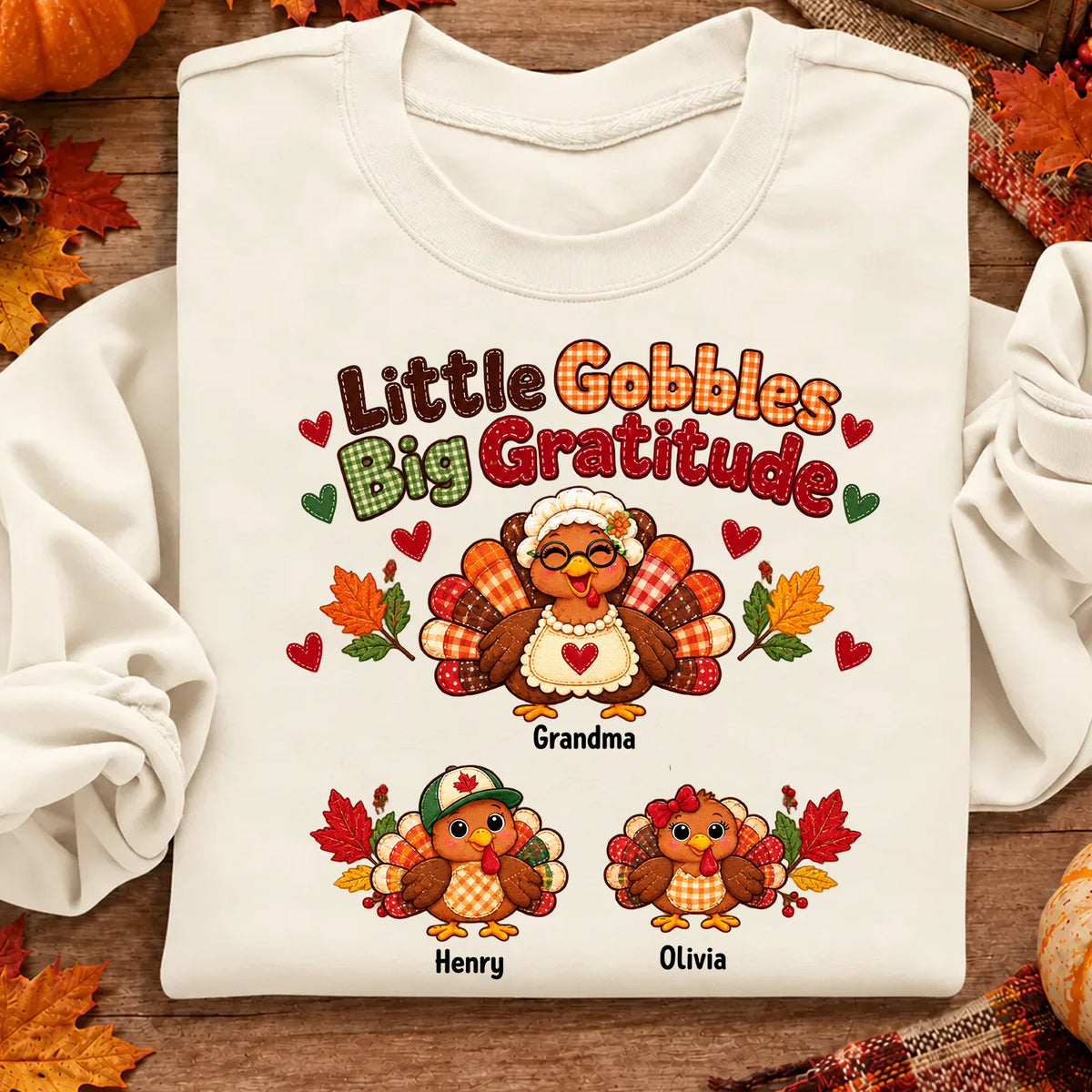 Personalized Little Gobbles Big Gratitude Sweatshirt in cream, folded flat with autumn leaves and plaid cloth around it