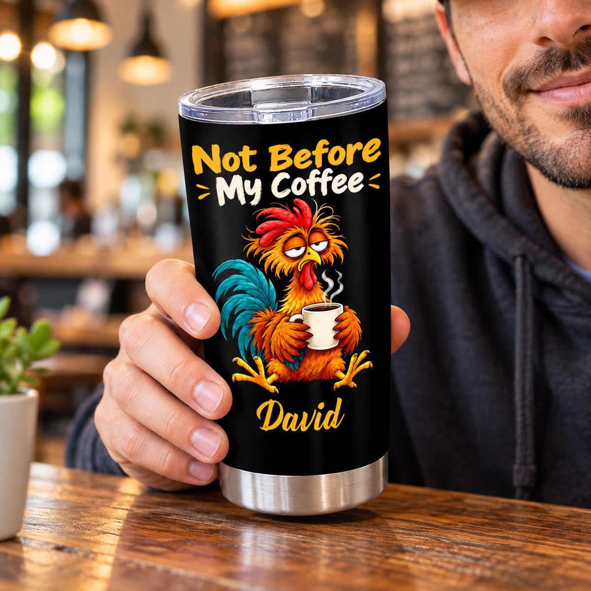 Personalized tumbler Not Before My Coffee held by a smiling man in a hoodie at a cafe table